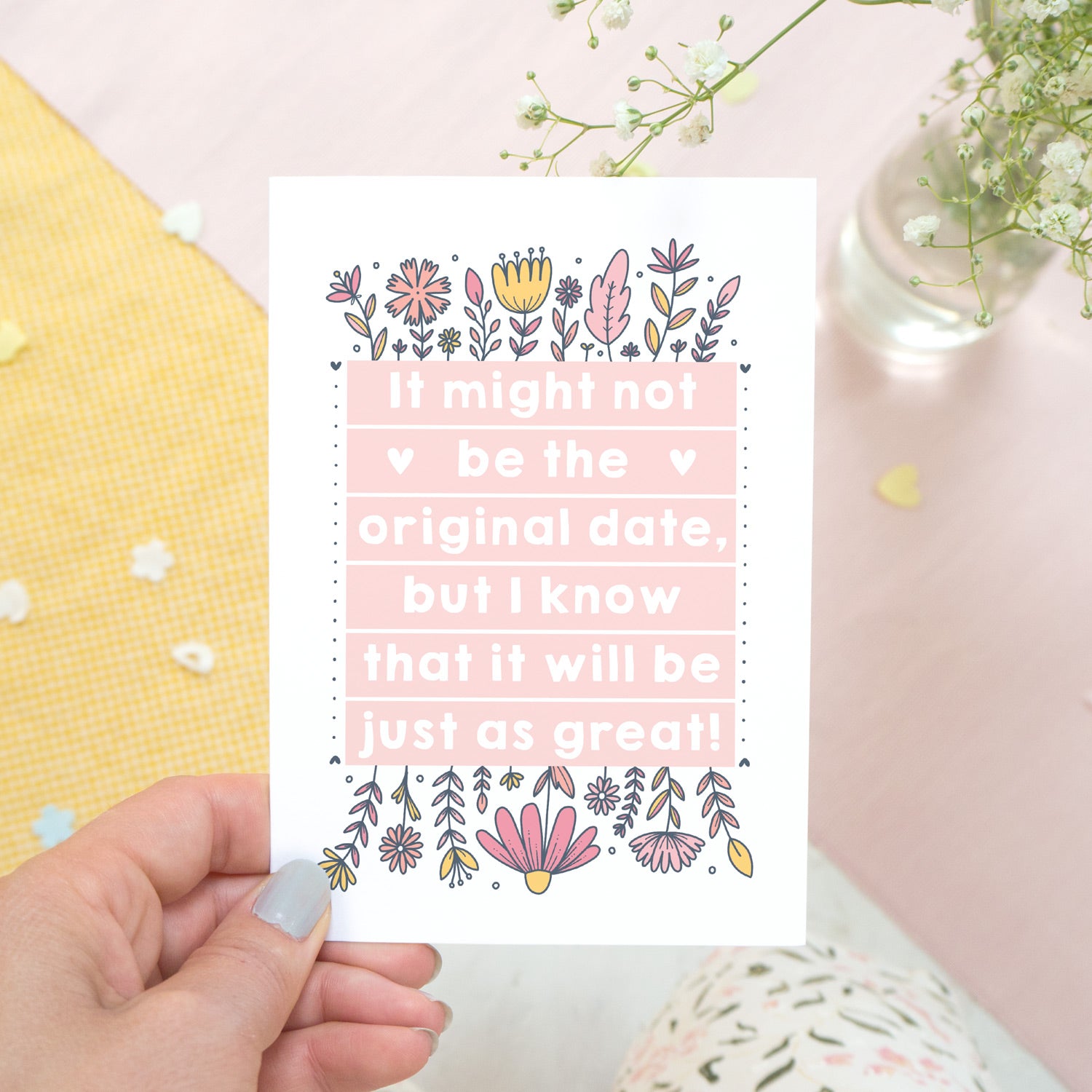 Original wedding date card for wedding postponements or delays. Photographed being held over a white, yellow and pink textured background with a pink pen and hint of foliage. The card features pink block of text and hand drawn florals.