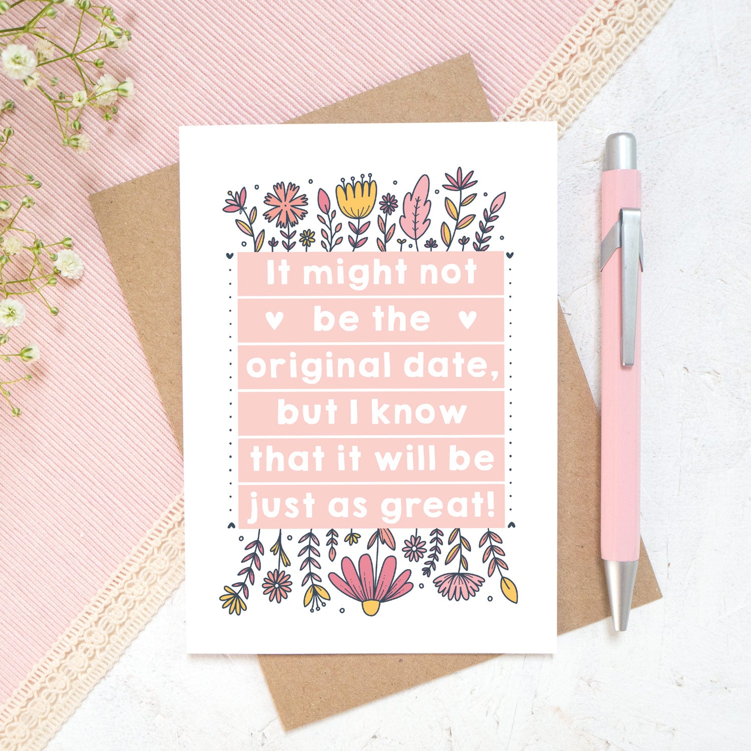 Original wedding date card for wedding postponements or delays. Photographed on a white and pink textured background with a pink pen and hint of foliage. The card features pink block of text and hand drawn florals.