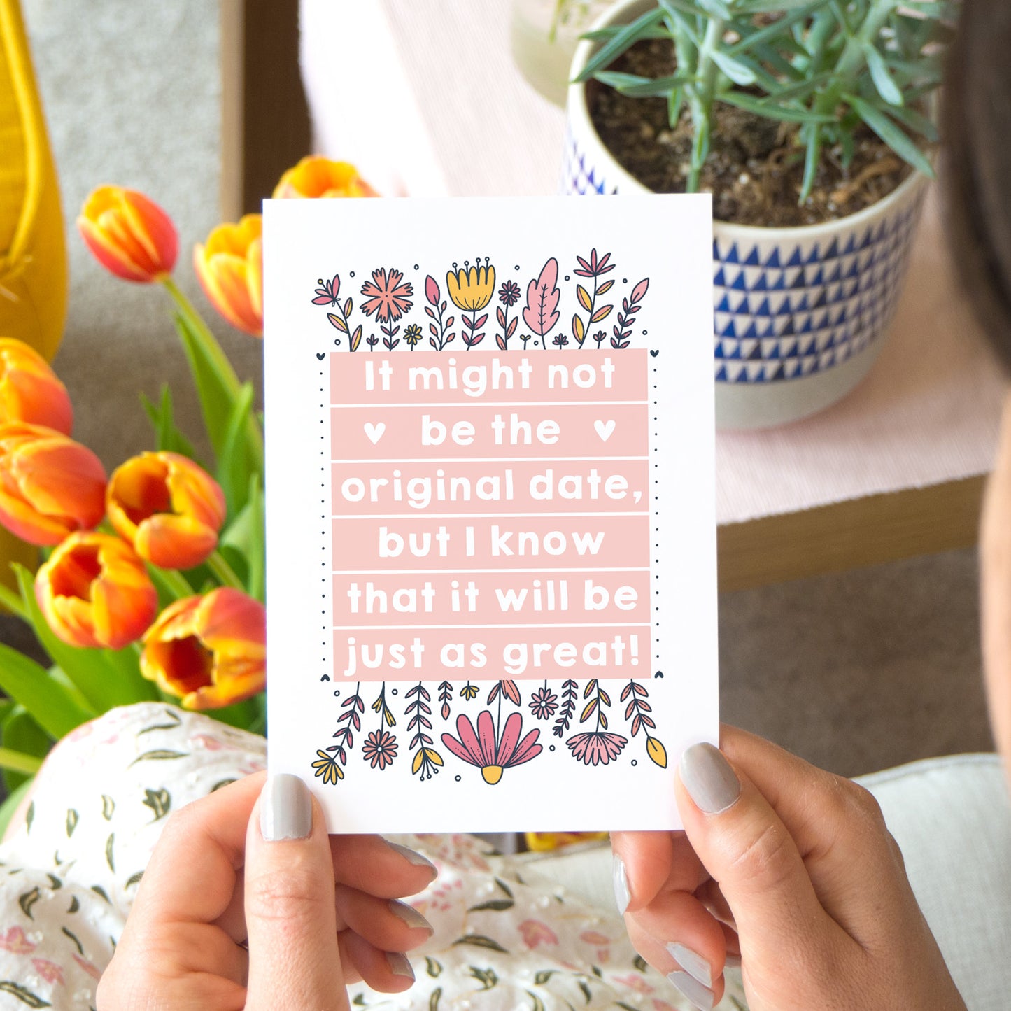 Original wedding date card for wedding postponements or delays. Photographed in a lifestyle setting with tulips, a person and plants on a table.. The card features pink block of text and hand drawn florals.