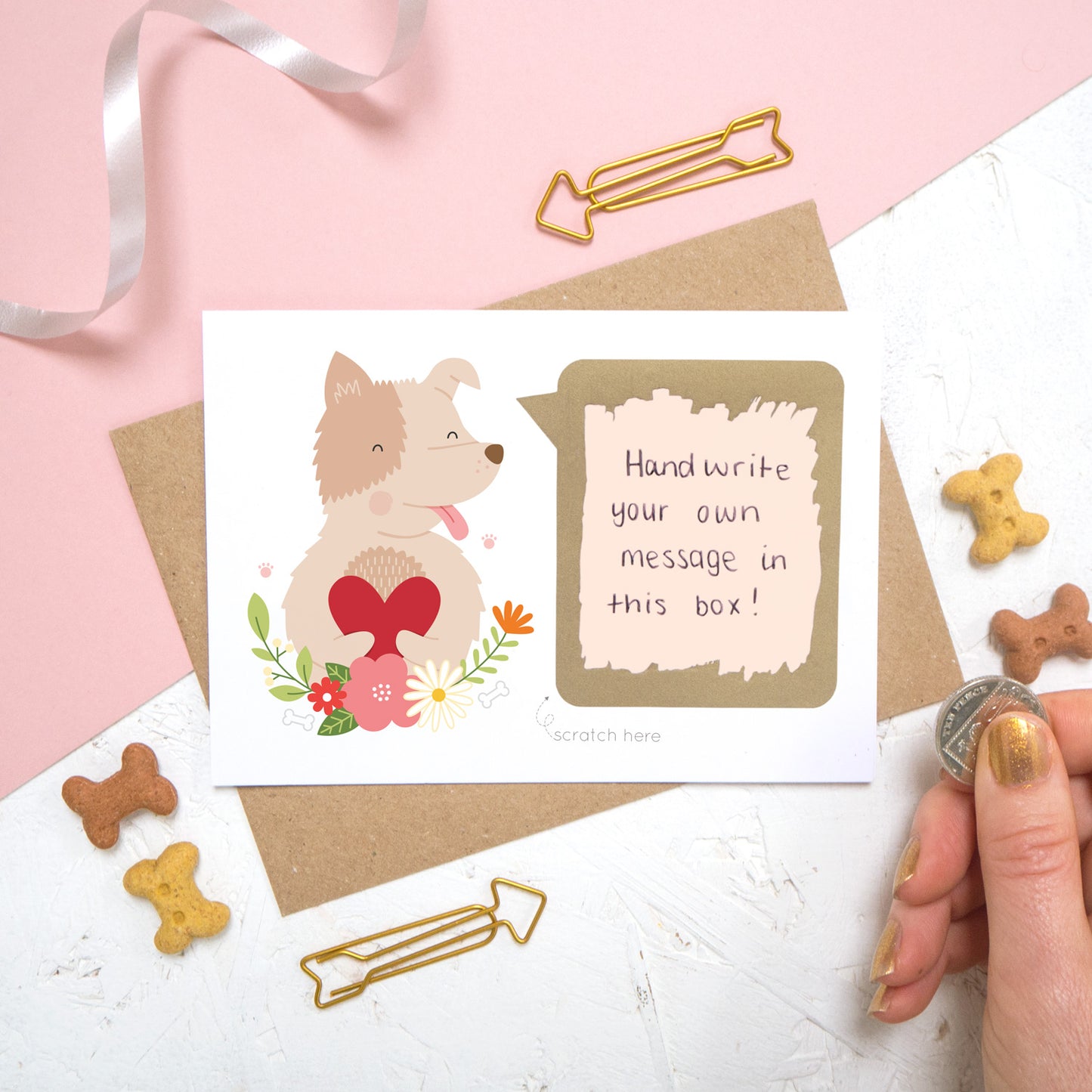 A personalised dog scratch card with a hand written message that has been scratched off. Photographed on a pink and white background with dog biscuits for props.