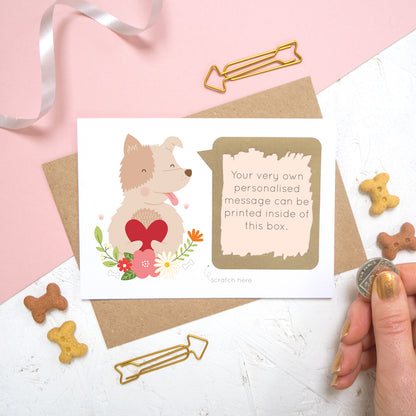 A personalised dog scratch card with a printed message that has been scratched off. Photographed on a pink and white background with dog biscuits for props.