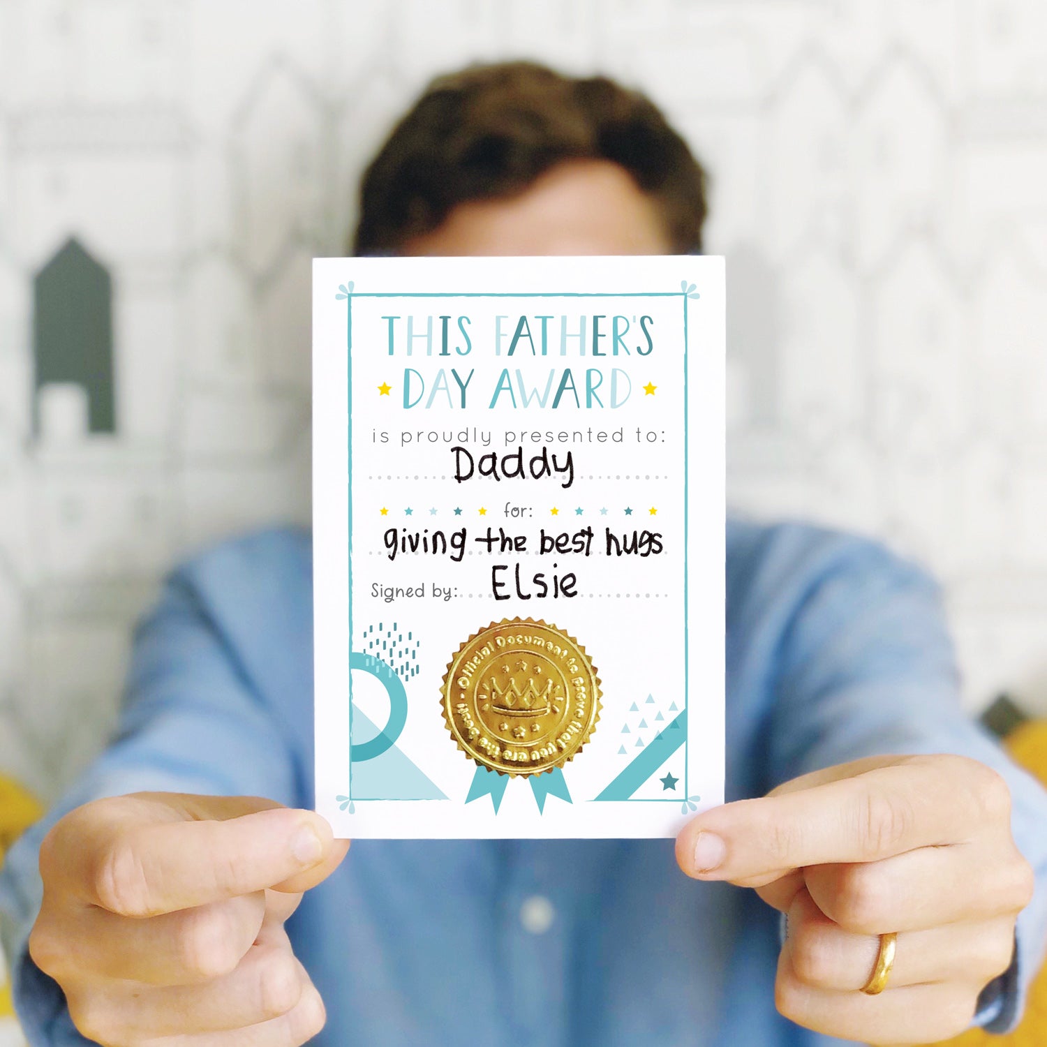A Father's day award card being held towards the camera by a male in a blue shirt. The card has been personalised by a childs handwriting and features a gold shiny certificate seal.