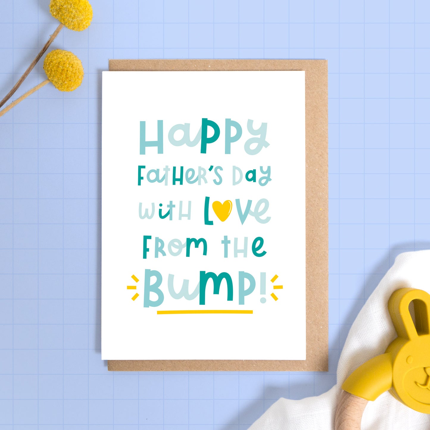 A happy father’s day from the baby bump card set on a blue background with some yellow flowers a muslin cloth and a baby soother.