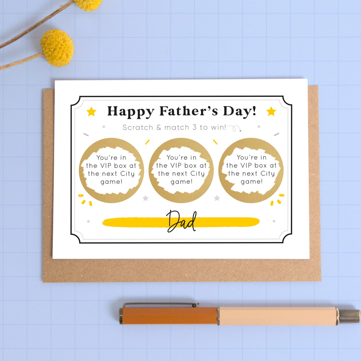 A personalised Father’s day scratch three to win style scratch card photographed on a blue background with a pop of yellow flowers and a pen for scale. This image shows the black and yellow version of the card with the golden circles scratched off to reveal the message.