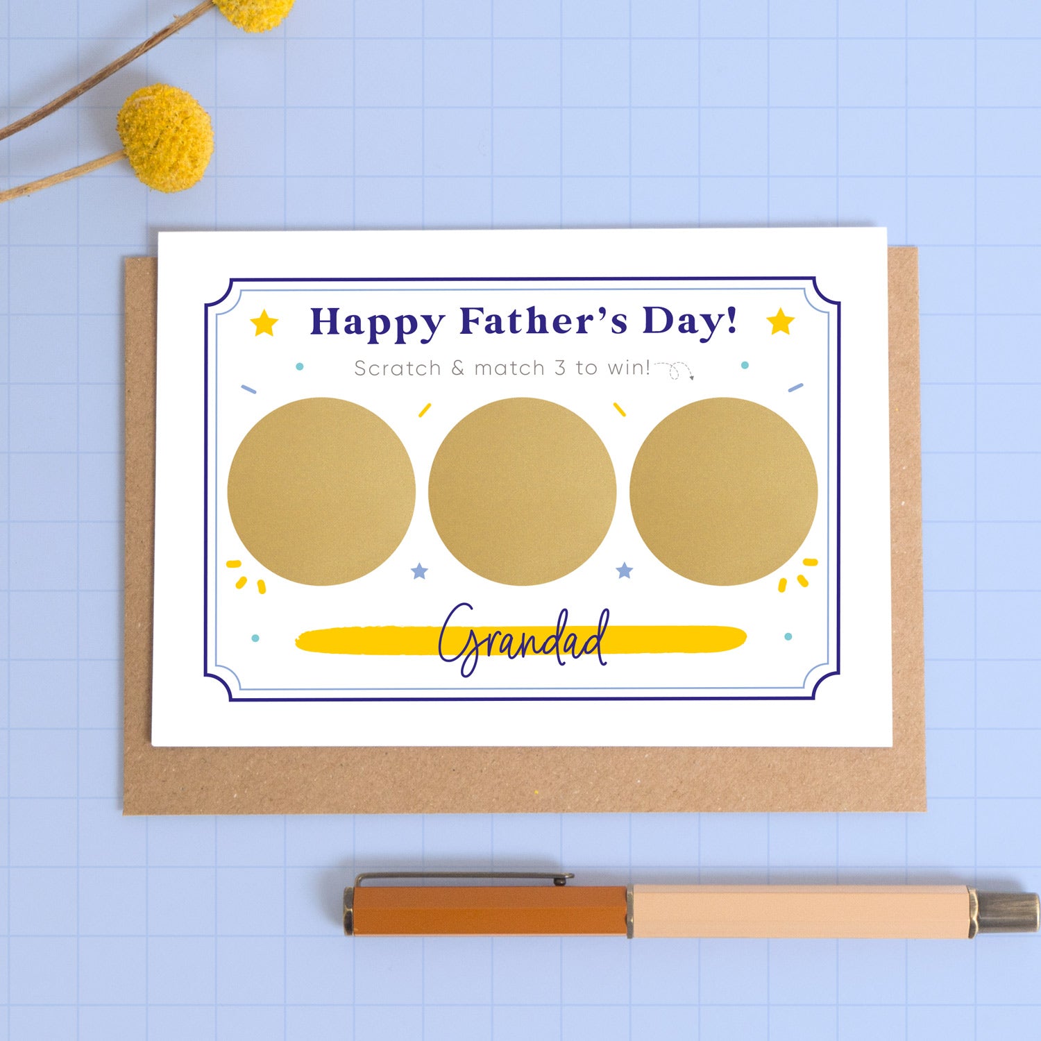 A personalised Father’s day scratch three to win style scratch card photographed on a blue background with a pop of yellow flowers and a pen for scale. This image shows the blue version of the card with the golden circles not yet scratched off.