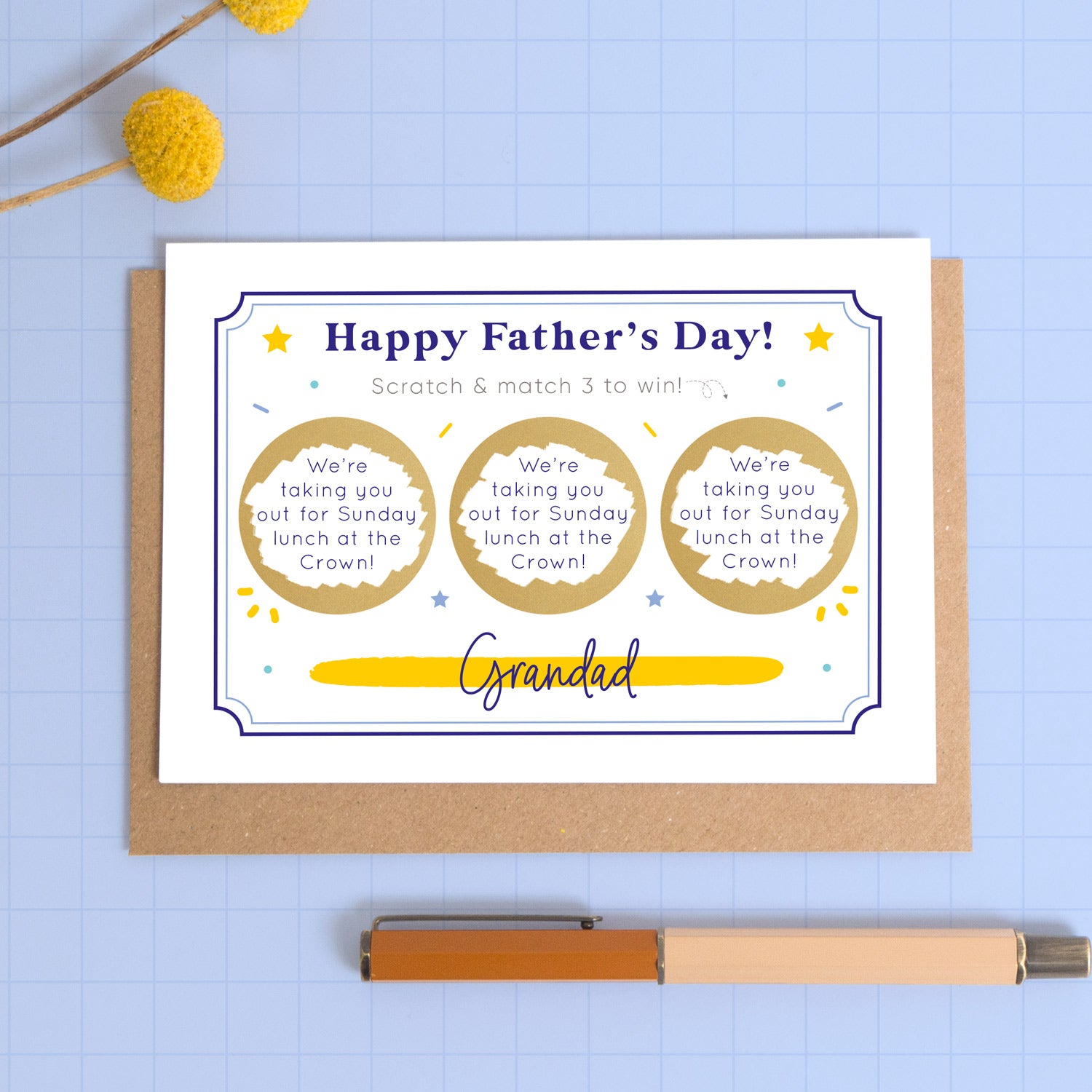A personalised Father’s day scratch three to win style scratch card photographed on a blue background with a pop of yellow flowers and a pen for scale. This image shows the blue version of the card with the golden circles scratched off to reveal the message.