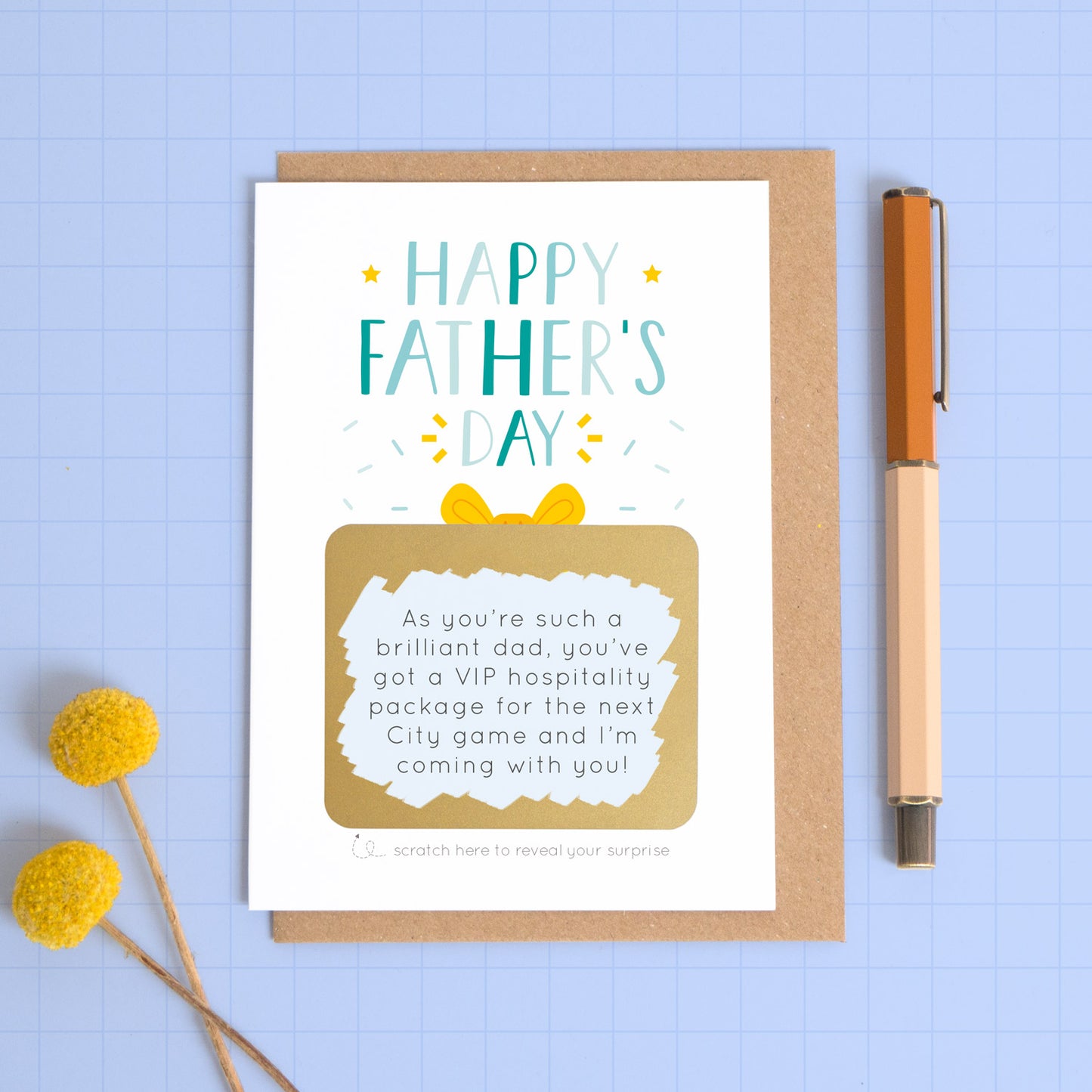 A personalised Father’s day scratch card photographed on a blue background with a pop of yellow flowers and a pen for scale. This image shows the blue version of the card with the golden present scratched off to reveal the message.