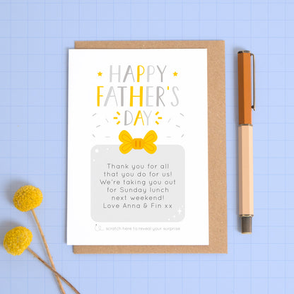A personalised Father’s day scratch card photographed on a blue background with a pop of yellow flowers and a pen for scale. This image shows the grey and yellow version of the card with the golden present fully scratched off to reveal the artwork under the scratch off box.