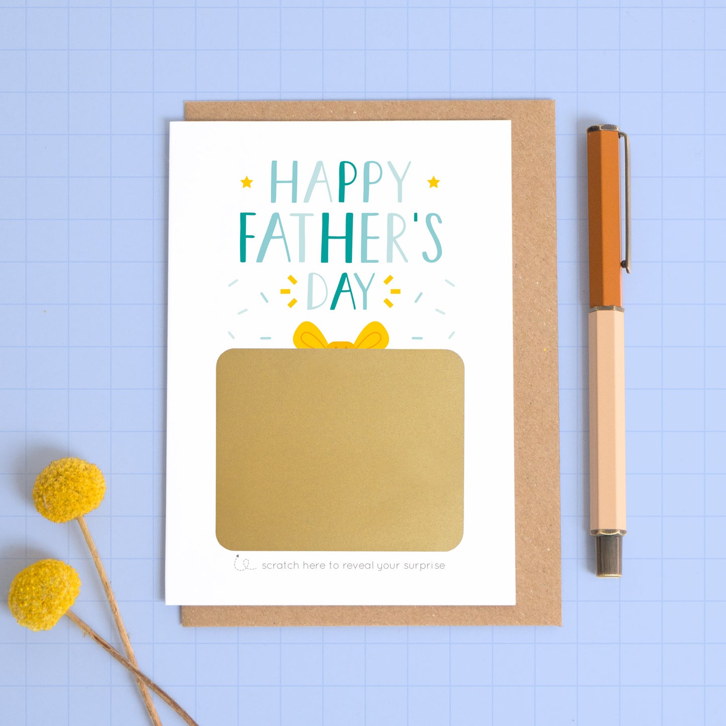 A personalised Father’s day scratch card photographed on a blue background with a pop of yellow flowers and a pen for scale. This image shows the blue version of the card with the golden present not yet scratched off. This is how the card will arrive.