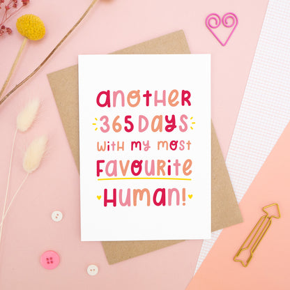 The 'another 365 days with my most favourite human' card photographed on a pink background with dried flowers, buttons and paper clips as props.