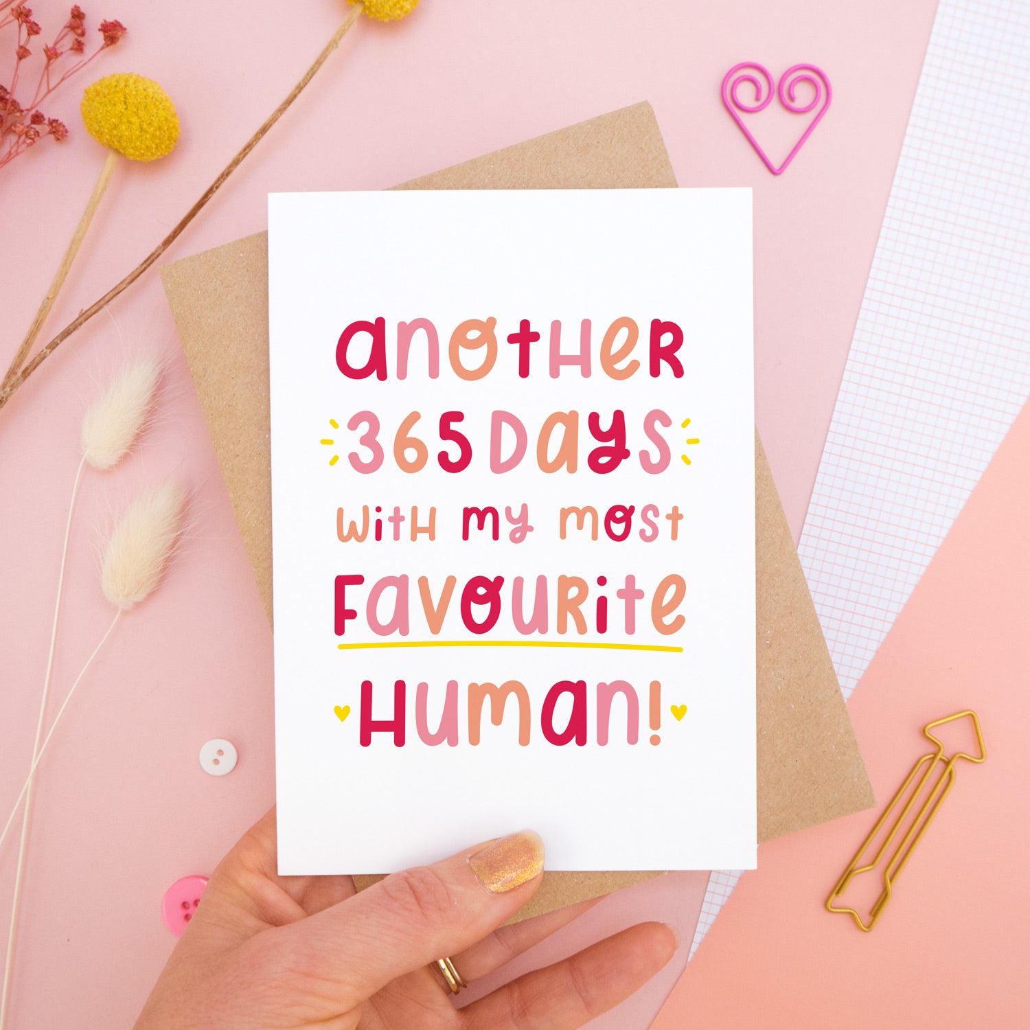 The 'another 365 days with my most favourite human' card photographed on a pink background with dried flowers, buttons and paper clips as props. The card itself is being held above the scene.