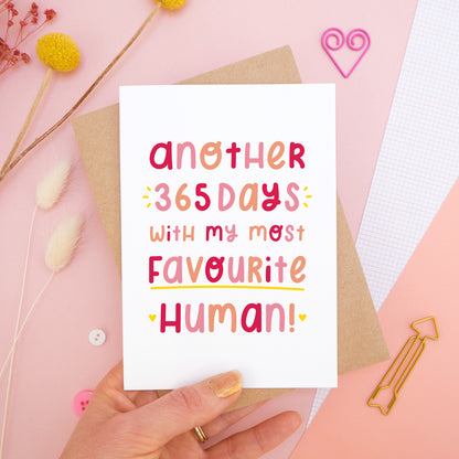The 'another 365 days with my most favourite human' card photographed on a pink background with dried flowers, buttons and paper clips as props. The card itself is being held above the scene.