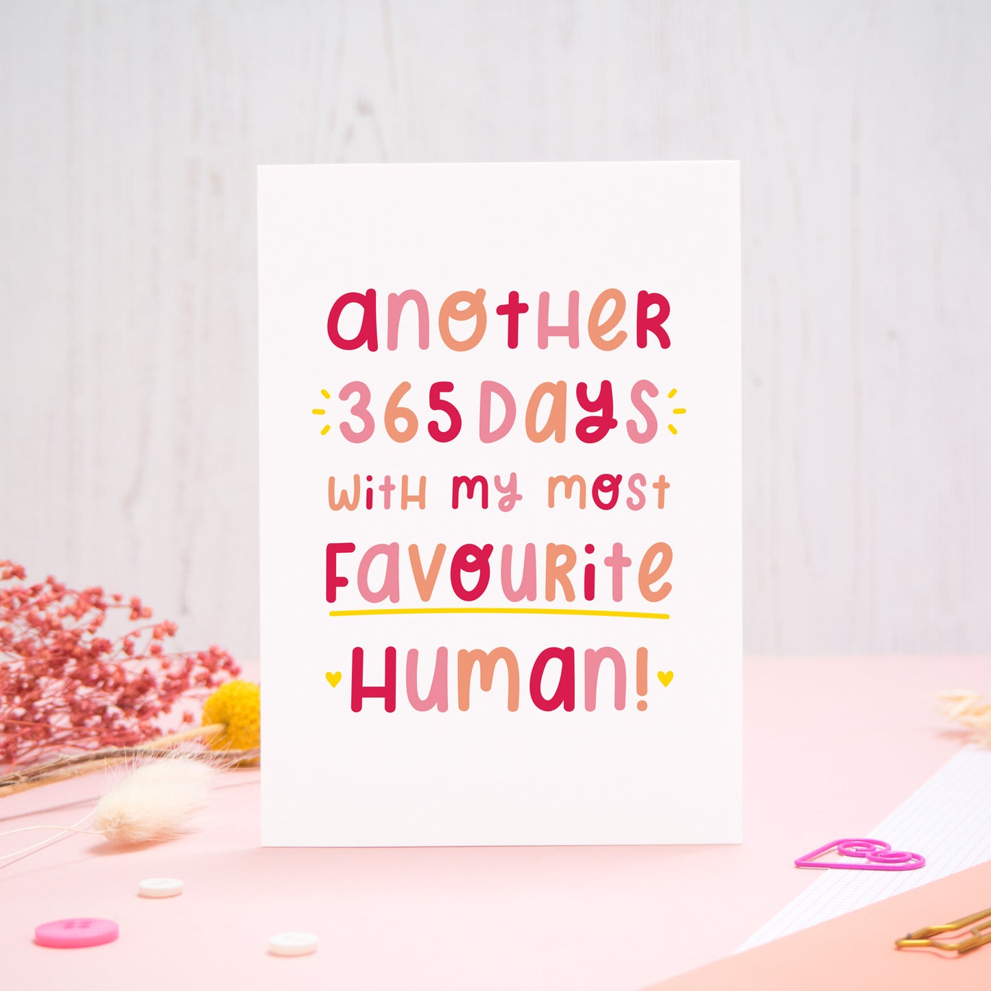 The 'another 365 days with my most favourite human' card photographed standing up against an off white background with flowers, buttons and paperclips at the base of the card.