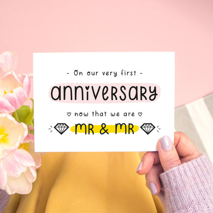 A first anniversary or Valentine’s card photographed on a pink background with pink tulip flowers, a grey and white stripe rug and yellow fabric. This image shows the first anniversary option with the Mr & Mr wording. The text is black and there are pops of yellow and pink behind key words.