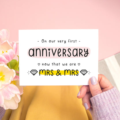 A first anniversary or Valentine’s card photographed on a pink background with pink tulip flowers, a grey and white stripe rug and yellow fabric. This image shows the first anniversary option with the Mrs & Mrs wording. The text is black and there are pops of yellow and pink behind key words.
