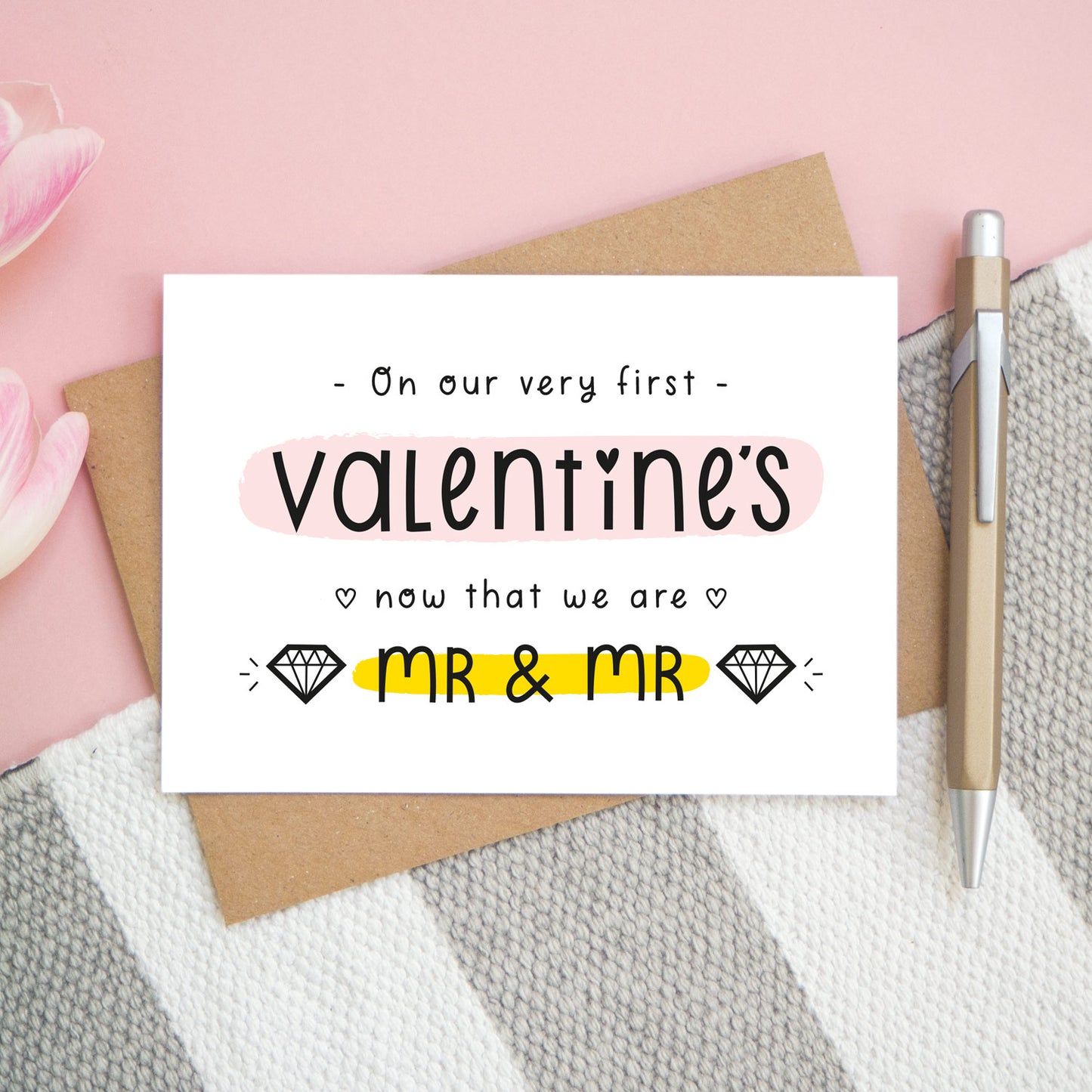 A first anniversary or Valentine’s card photographed on a pink background with pink tulip flowers, a gold pen and a grey and white stripe rug. This image shows the first valentine’s option with the Mr & Mr wording. The text is black and there are pops of yellow and pink behind key words.