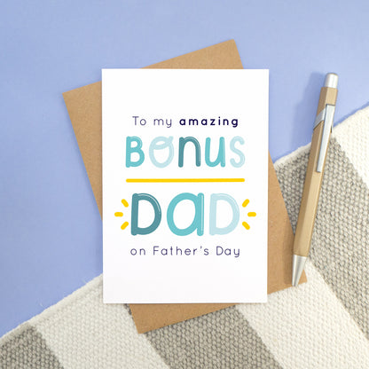 A bonus dad card laying on a stripy carpet and a blue background. The card features navy and varying tones of blue wording and a yellow underline and flicks.