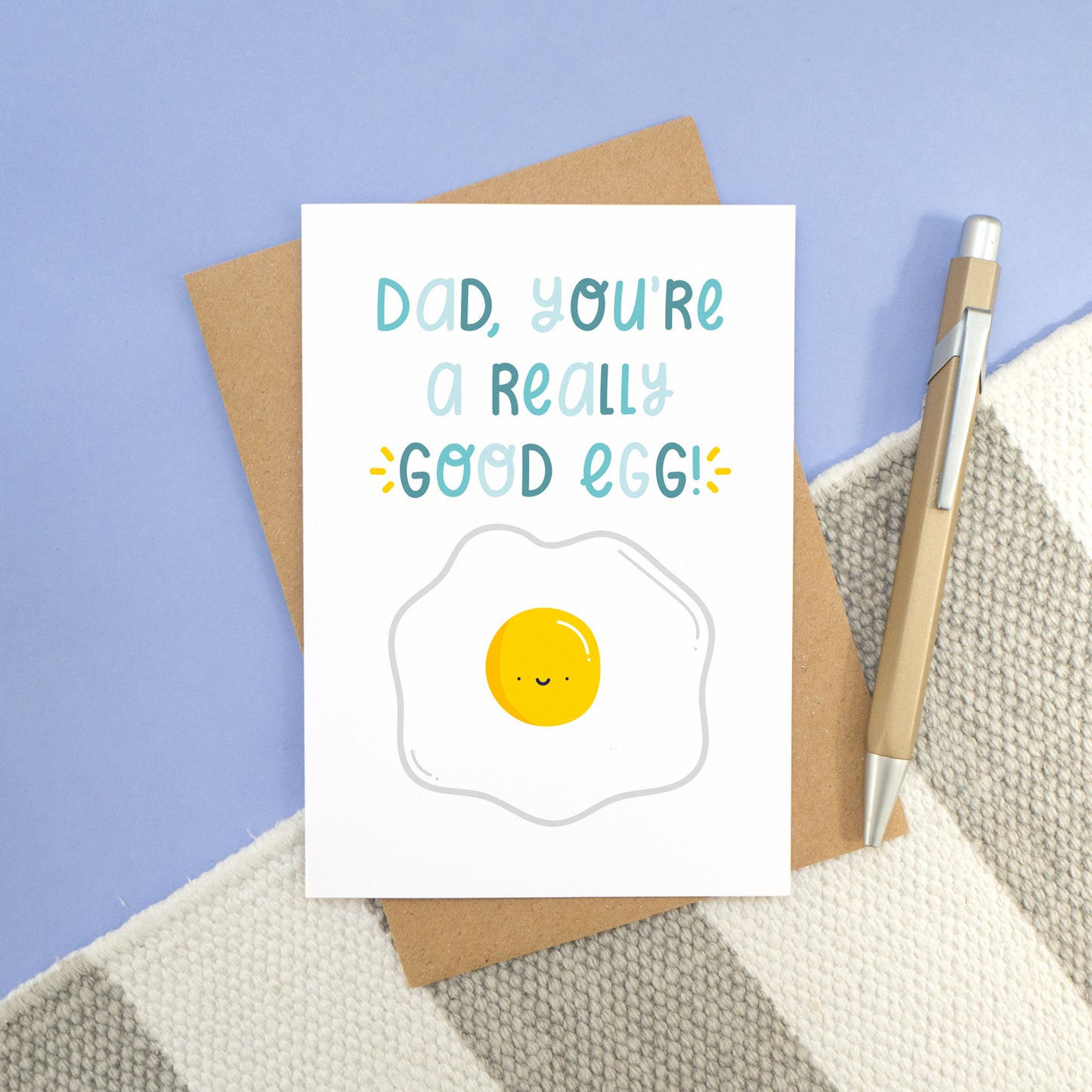A good egg card photographed on a stripy white and grey rug, with a blue background and a pen. The card features text in varying tones of blue and a smiley happy egg!