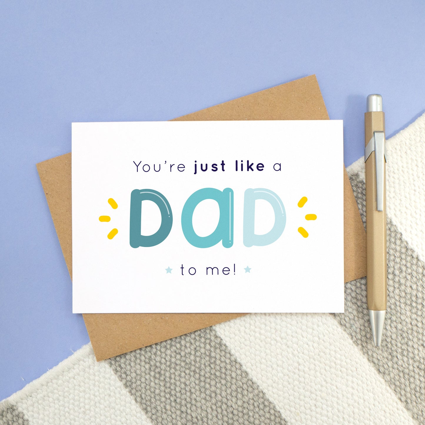 A 'like a dad to me' card shot on top of a kraft envelope and stripy rug with a blue background with a pen. The card shows the text 'you're just like a dad to me!' in varying tones of blue and navy with yellow flicks.