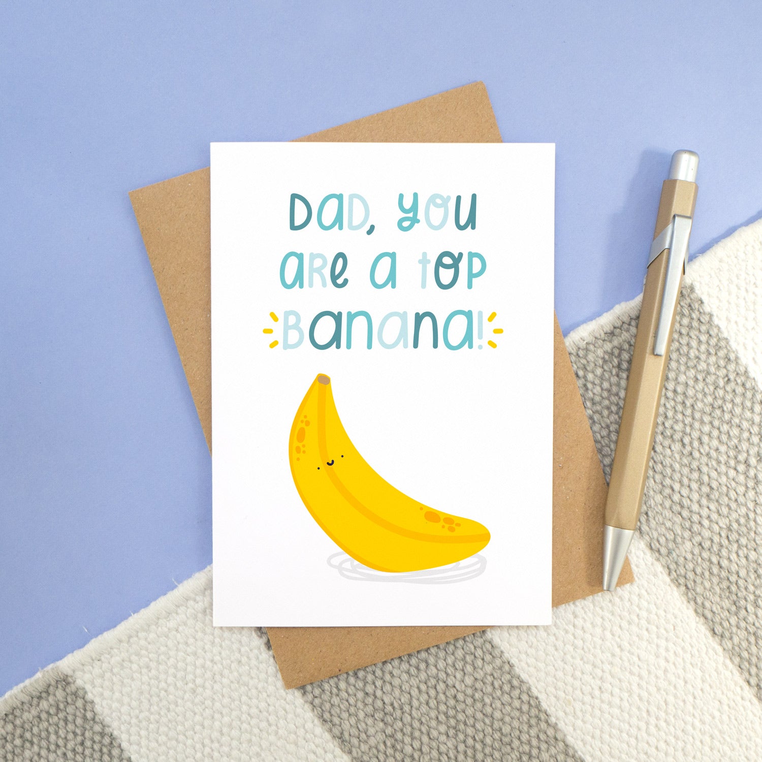 A top banana card photographed on a stripy white and grey rug, with a blue background and a pen. The card is lying on a kraft brown envelope and feature blue lettering with a happy bright yellow banana!