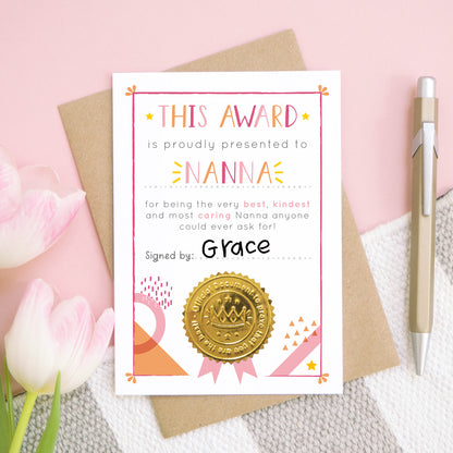 A Nanna certificate card for mother's day featuring a shiny gold seal. This card is pink and peach in colour with a small pop of yellow and has been shot over head on a pink and grey and white background. There is a gold pen for scale and tulips on the left.