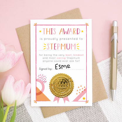A stepmum certificate card for mother's day featuring a shiny gold seal. This card is pink and peach in colour with a small pop of yellow and has been shot over head on a pink and grey and white background. There is a gold pen for scale and tulips on the left.