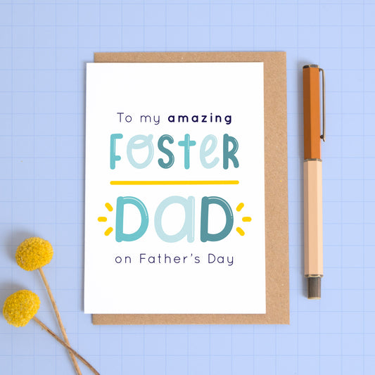 A foster dad Father’s day card photographed on a blue background with a pop of yellow flowers and a pen for scale. The card reads “to my amazing foster dad on Father’s day”.