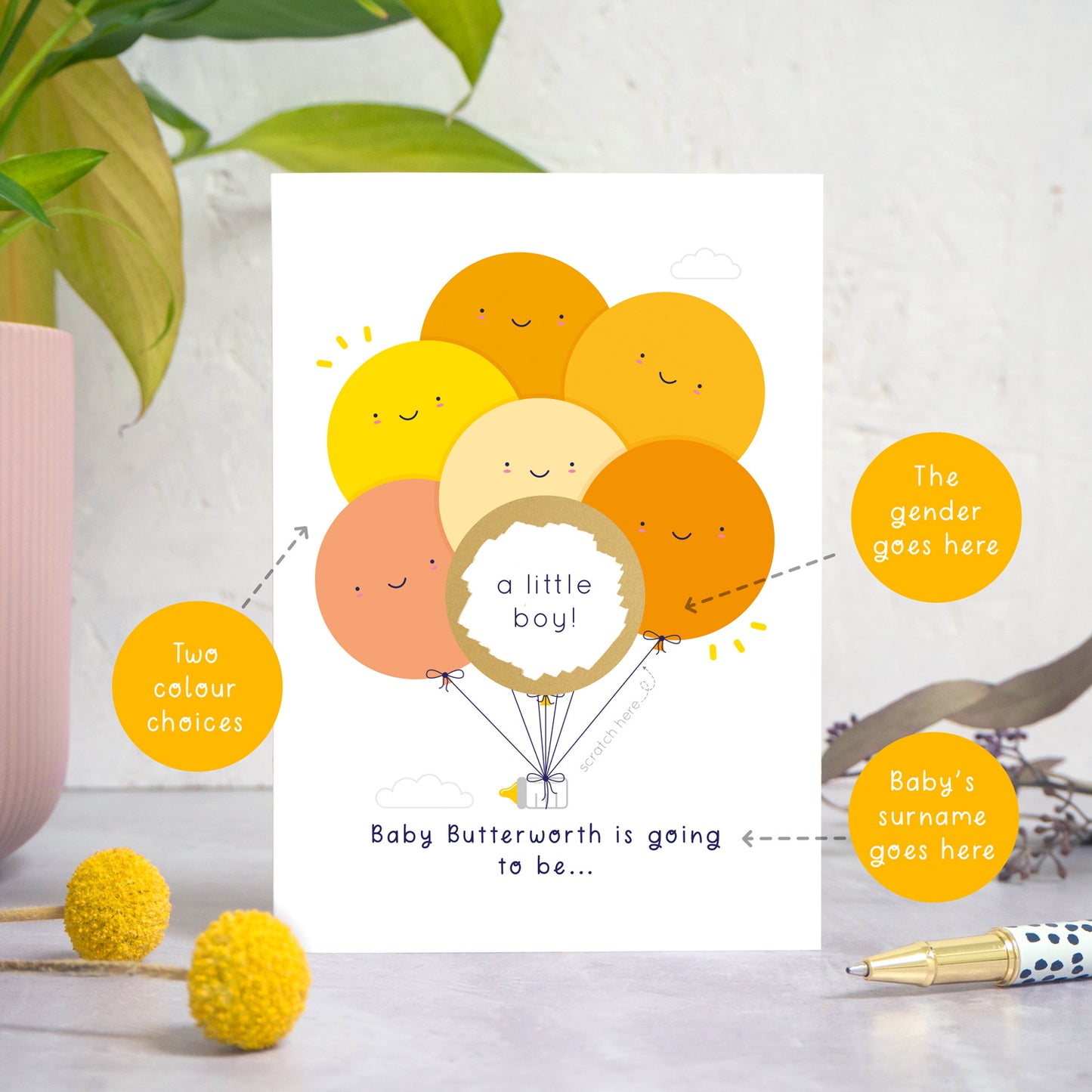 A ‘gender reveal’ scratch card photographed on a white and grey background with a plant on the left and foliage and a pen on the right. This image demonstrates the custom options of the card, eg. the colour of the balloons, the gender and the baby’s surname.