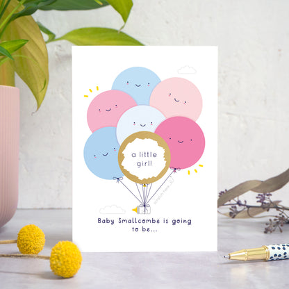 A ‘gender reveal’ scratch card photographed on a white and grey background with a plant on the left and foliage and a pen on the right. This is the pink & blue version of the card after it has been scratched off.