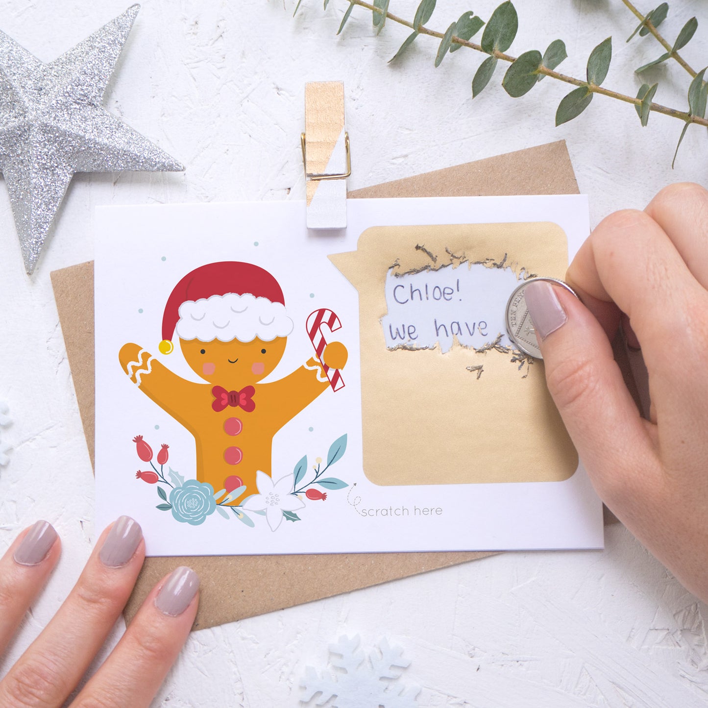 A personalised gingerbread man scratch card where the scratching off of the gold panel is being demonstrated. Shot on a white background with a glittery star and sprig of eucalyptus.