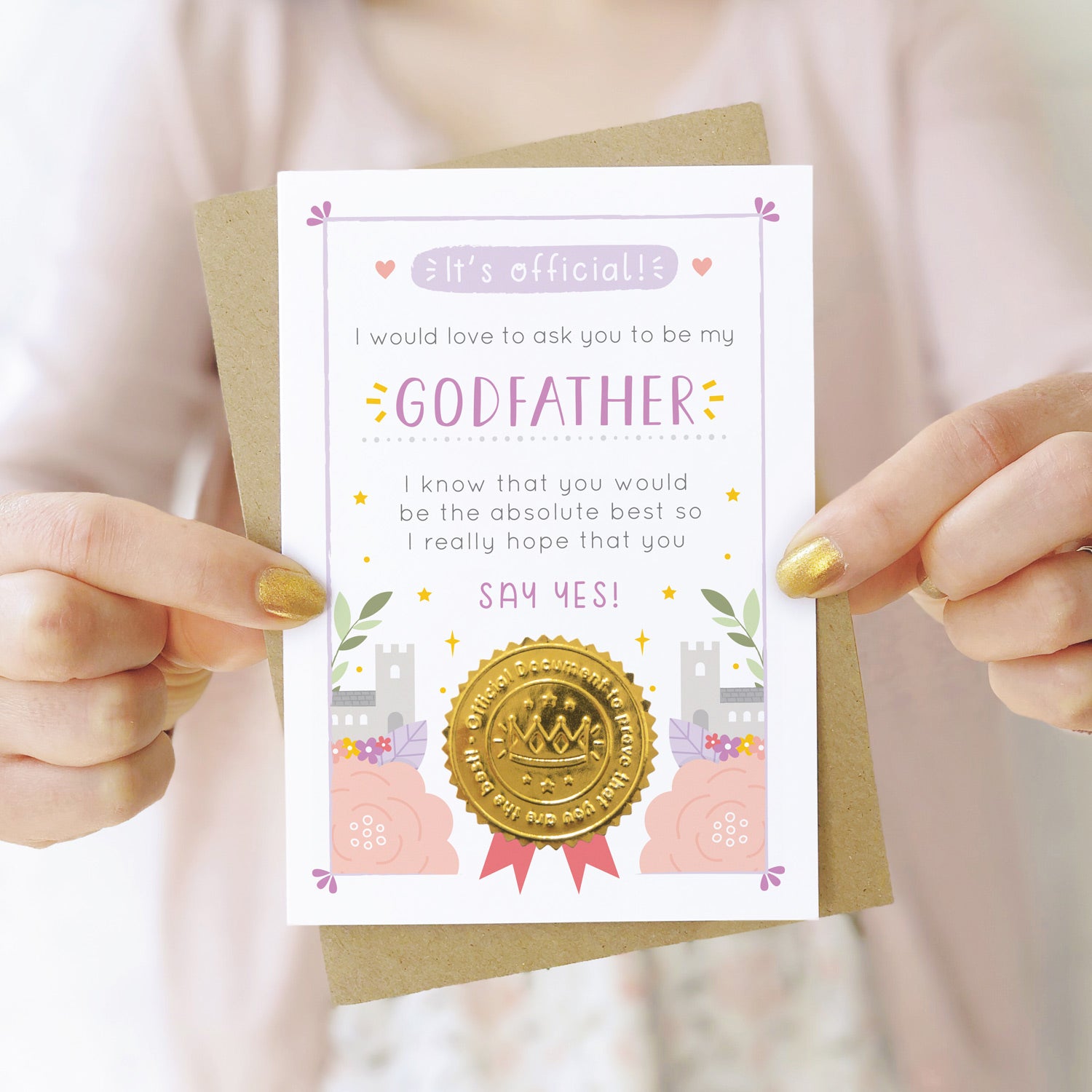 A will you be my godfather card in purple and pink being held by both hands in front of a person wearing a pink cardigan and a white dress.