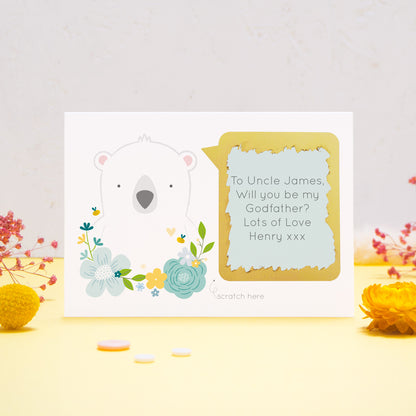 A personalised will you be my godfather scratch card featuring a white bear, blue flowers and a blue text box. This has been shot on a yellow and grey background with flowers. The gold panel has been scratched away to reveal the message.