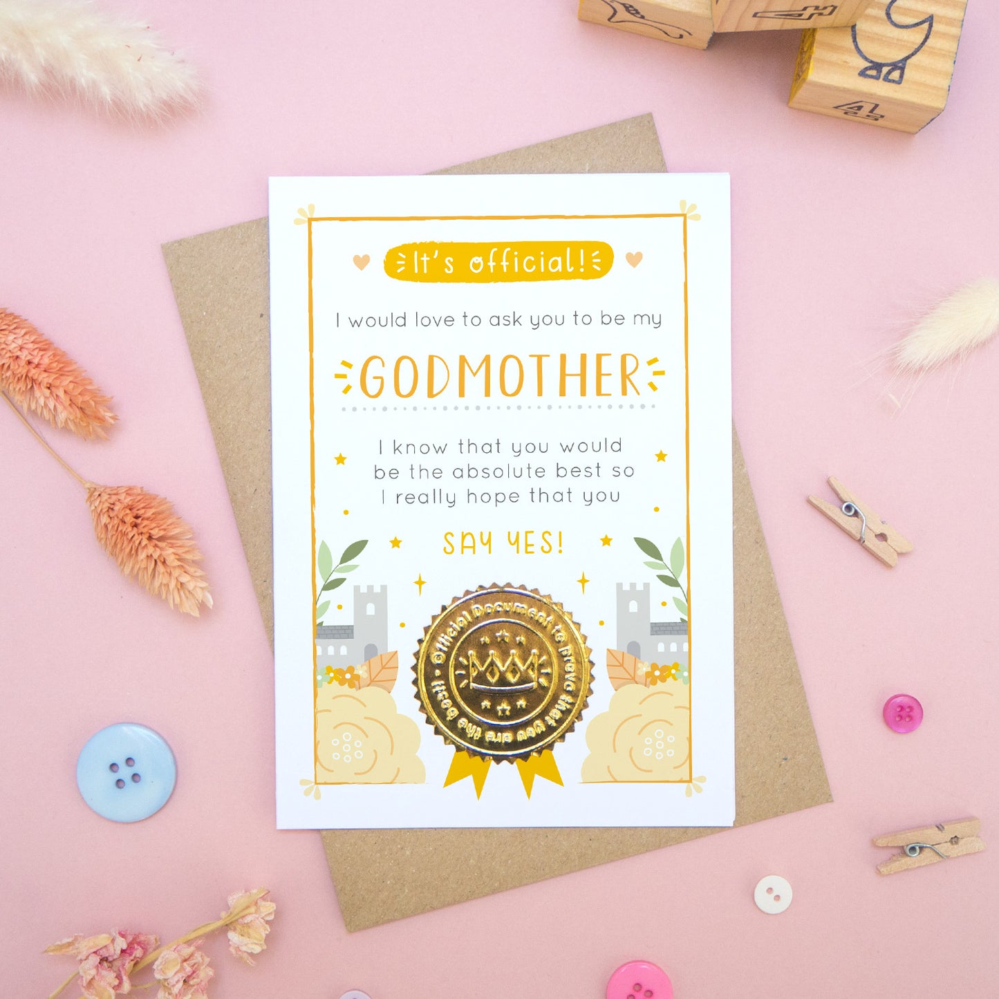 A will you be my godmother card in orange, shot on a pink background surrounded by dry flowers, buttons and building blocks.