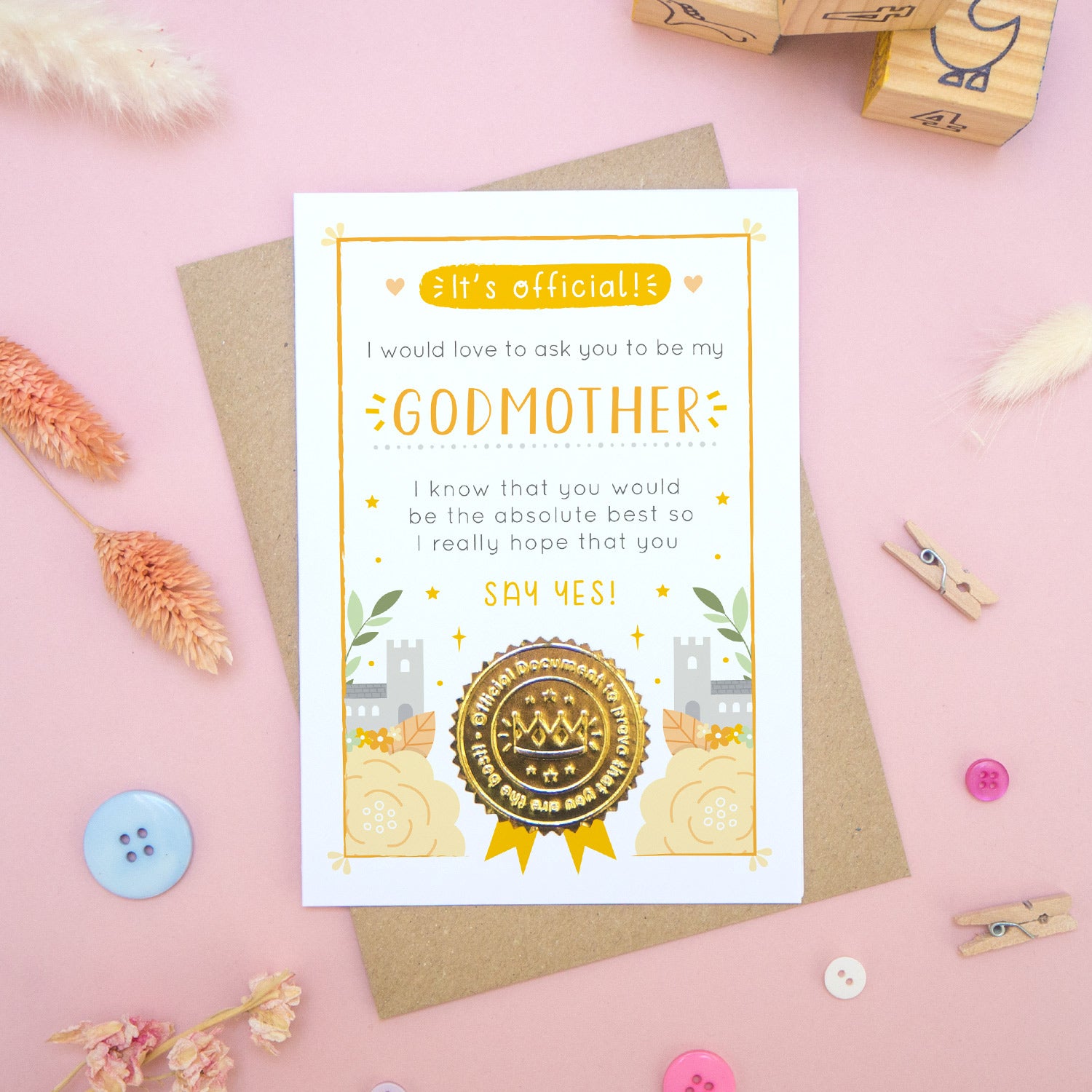 A will you be my godmother card in orange, shot on a pink background surrounded by dry flowers, buttons and building blocks.
