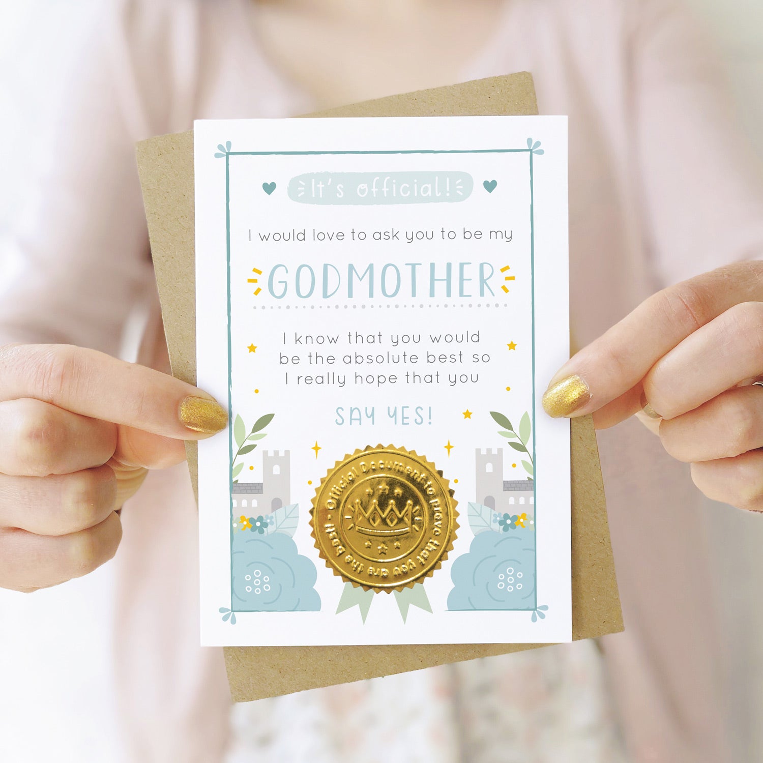 A will you be my godmother card in blue being held by both hands in front of a person wearing a pink cardigan and a white dress.