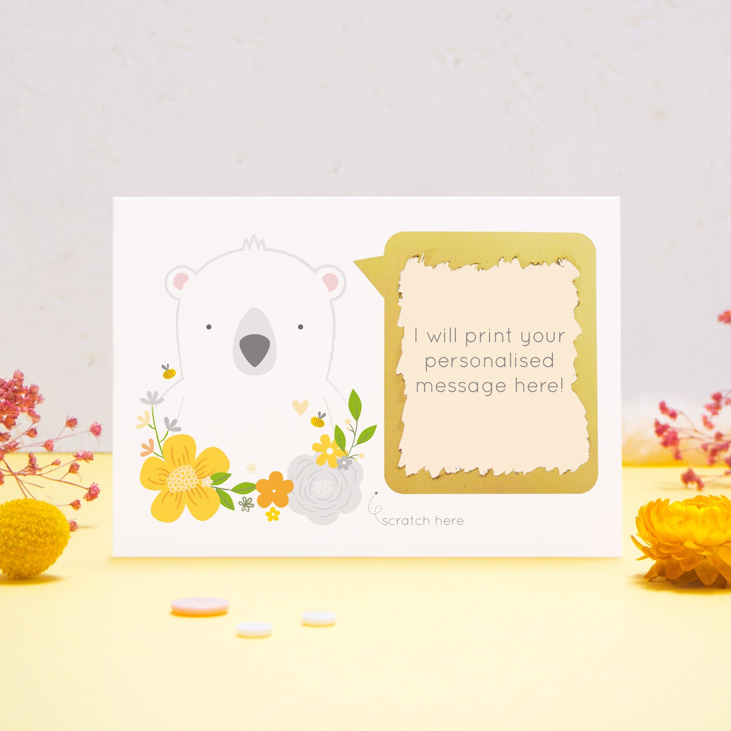A personalised scratch card showing where your message will be printed featuring a white bear, yellow flowers and a peach text box. This has been shot on a yellow and grey background with flowers. The gold panel has been scratched away to reveal the message.