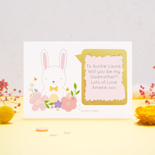 A personalised will you be my godmother scratch card featuring a white white, pink flowers and a pink text box. This has been shot on a yellow and grey background with flowers. The gold panel has been scratched away to reveal the message.