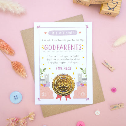 A will you be my godparents card in pink and purple, shot on a pink background surrounded by dry flowers, buttons and building blocks.