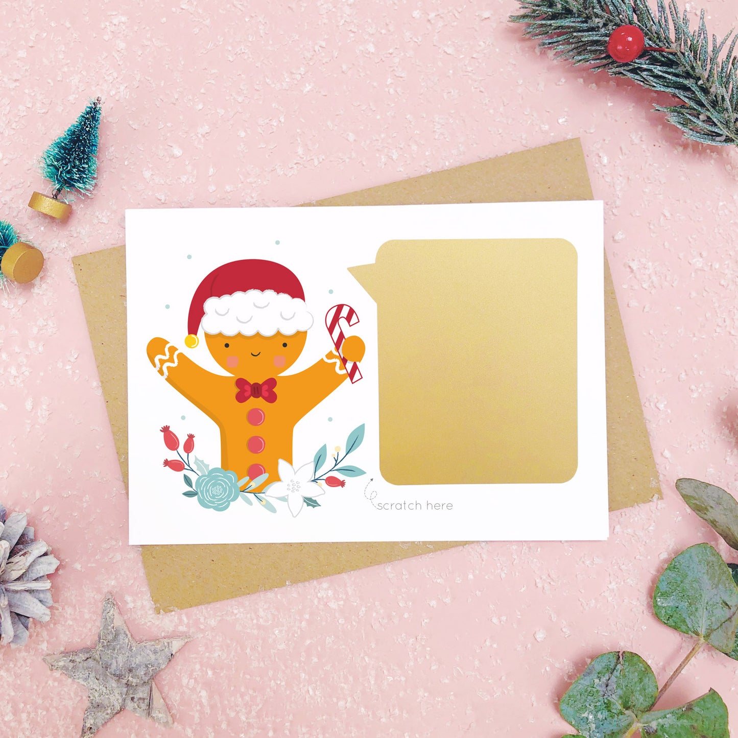 A personalised gingerbread man scratch card after the gold panel has been attached.Shot on a pink background with grey and green festive props.