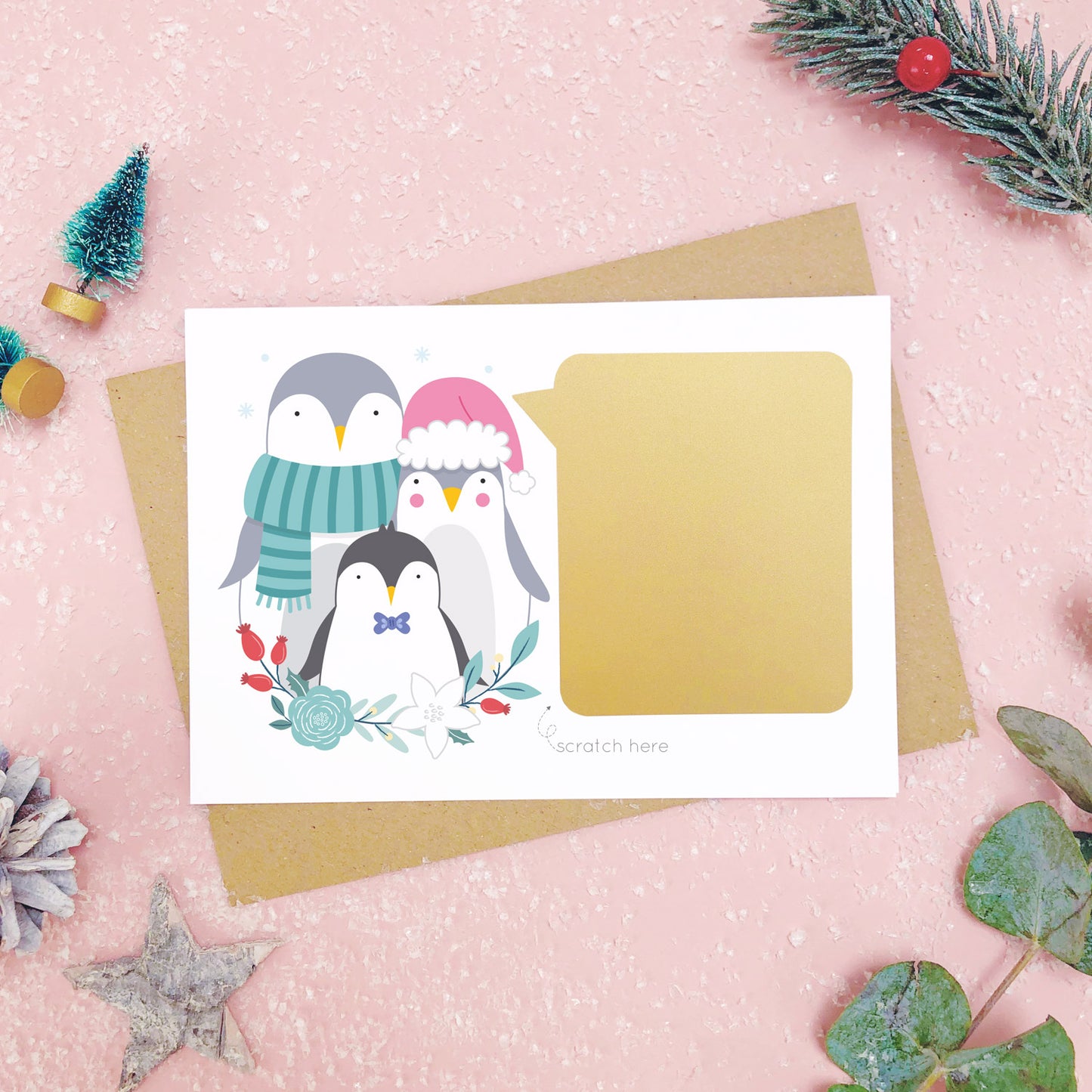 A personalised penguin family scratch card after the gold panel has been attached.Shot on a pink background with grey and green festive props.