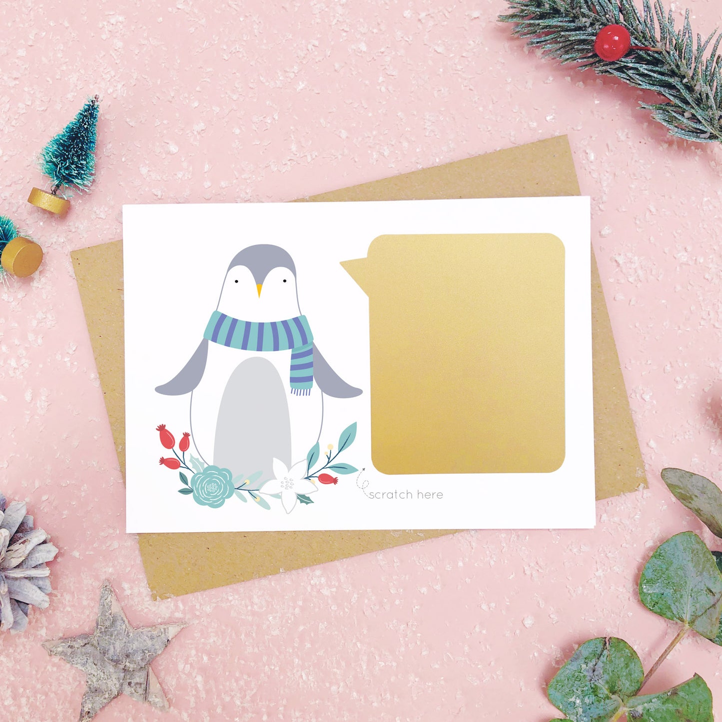 Penguin scratch card showing the gold panel after it has been stuck down. Shot on a pink background, surrounded with festive christmas props in tones of green and grey.