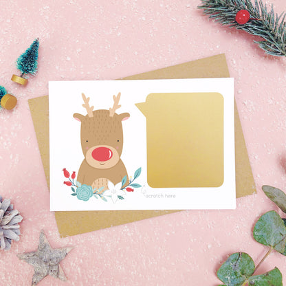A personalised red nose reindeer scratch card after the gold panel has been attached.Shot on a pink background with grey and green festive props.