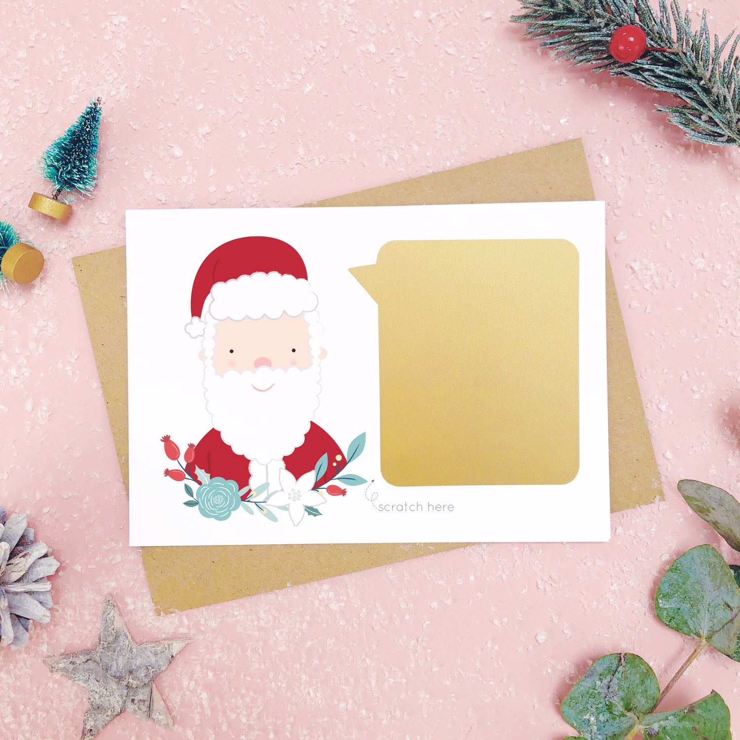 A santa scratch card with the gold panel attached to the front before it is scratched off. Shot on a pink background with green and grey festive props.