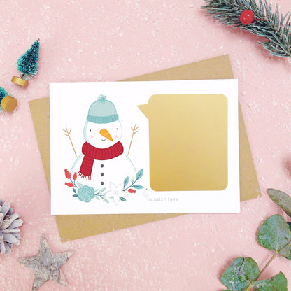 A personalised snowman scratch card after the gold panel has been attached.Shot on a pink background with grey and green festive props.