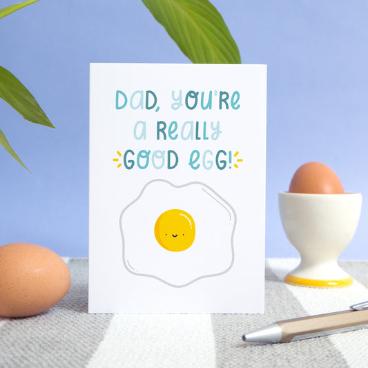 A good egg card photographed on a grey and white rug with a blue background, leaves, two eggs and a pen! The card features text in varying tones of blue and a smiley happy egg!