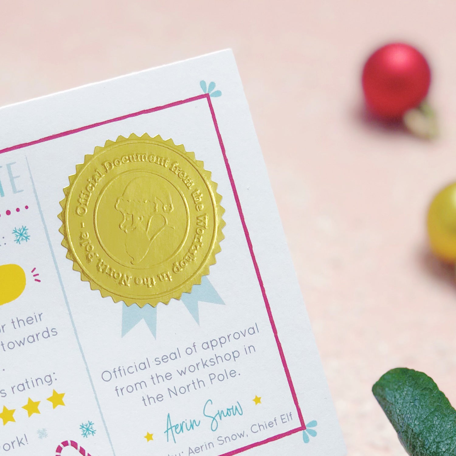 A close up of the shiny gold Christmas stamp. The stamp reads "Official document from the workshop in the north pole". Shot over a pink background with baubles and green foliage.