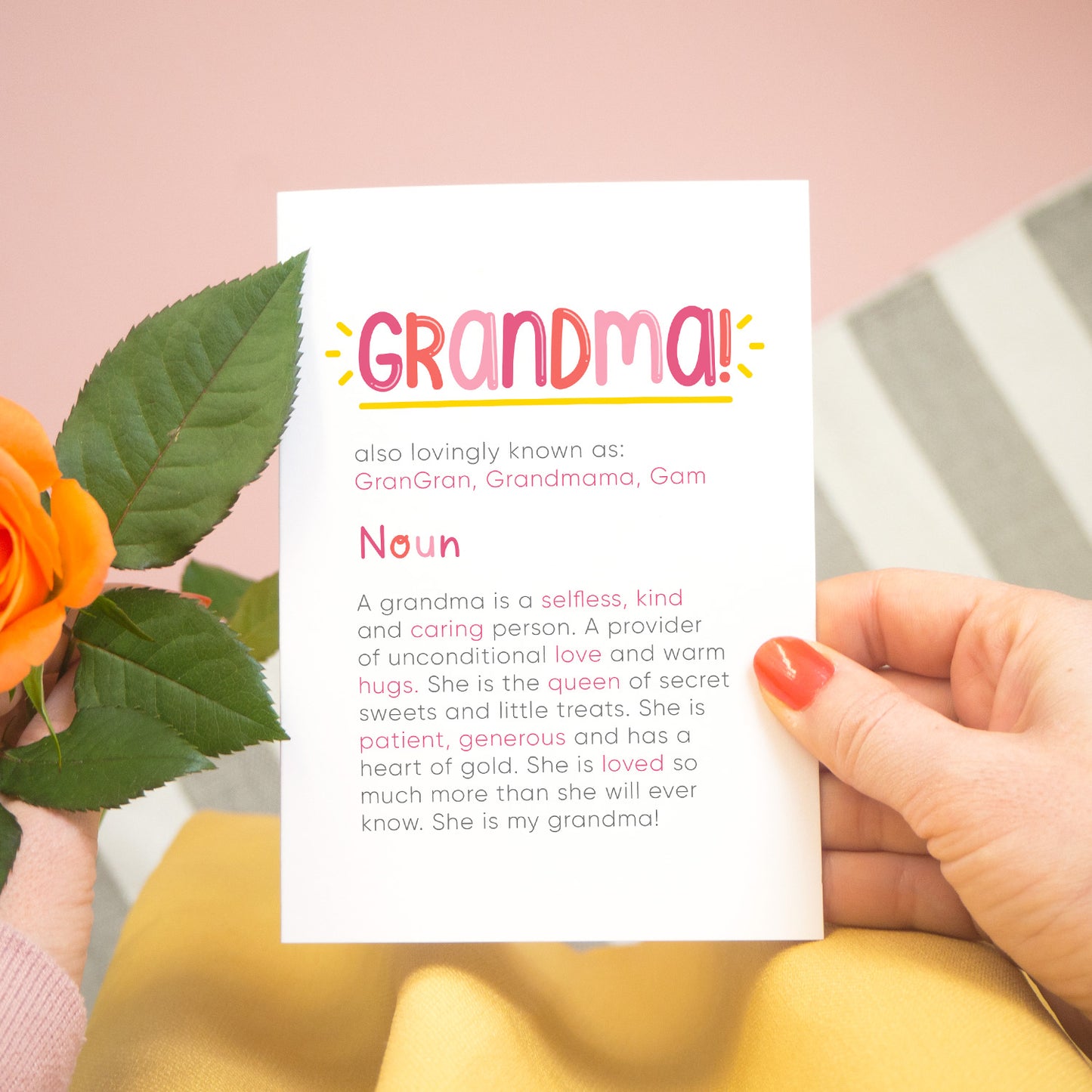 A personalised Grandma dictionary definition card being held over a pink background with a grey and white striped rug, yellow skirt and a single orange rose to the left. The card features hand drawn writing in varying tones of pink and a definition of what a Grandma is with key words highlighted in pink.