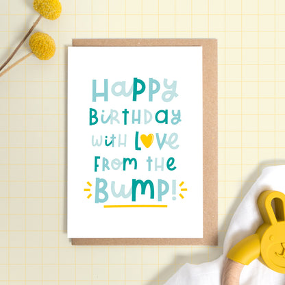 A happy birthday from the baby bump card set on a yellow background with some yellow flowers a muslin cloth and a baby soother. This is the version of the card in blue.