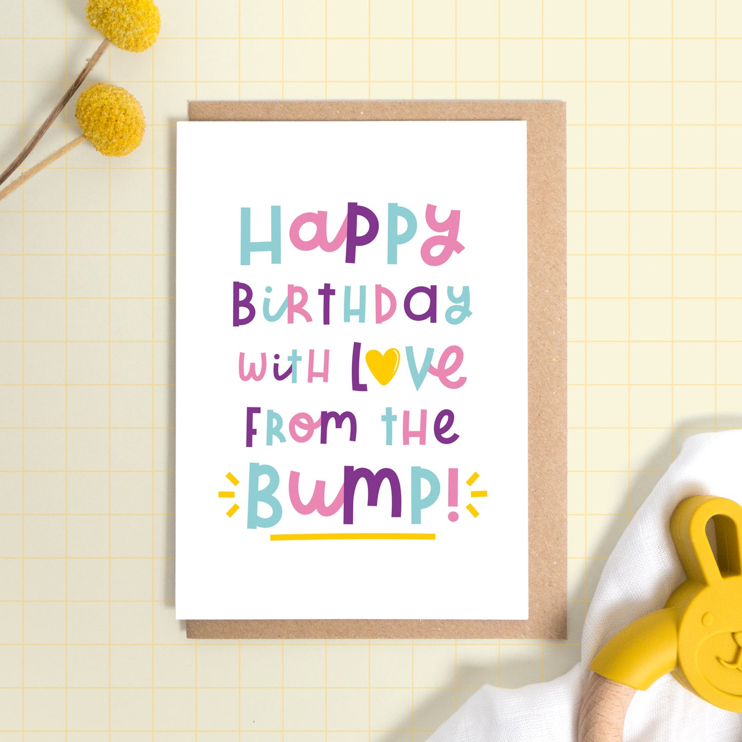 A happy birthday from the baby bump card set on a yellow background with some yellow flowers a muslin cloth and a baby soother. This is the version of the card in pink, purple and blue.