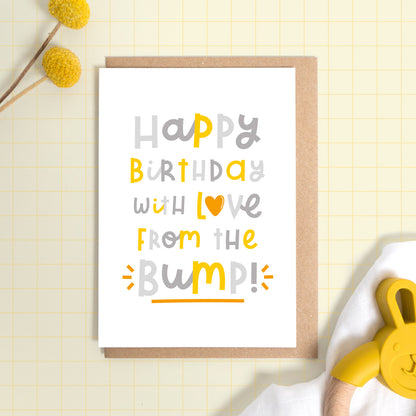 A happy birthday from the baby bump card set on a yellow background with some yellow flowers a muslin cloth and a baby soother. This is the version of the card in grey and yellow.